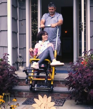 woman sitting on yellow framed wheelchair with track on of wheelchair. She is being assisted to ascend steps into a house by a man, stabilizing the handle of the wheelchair