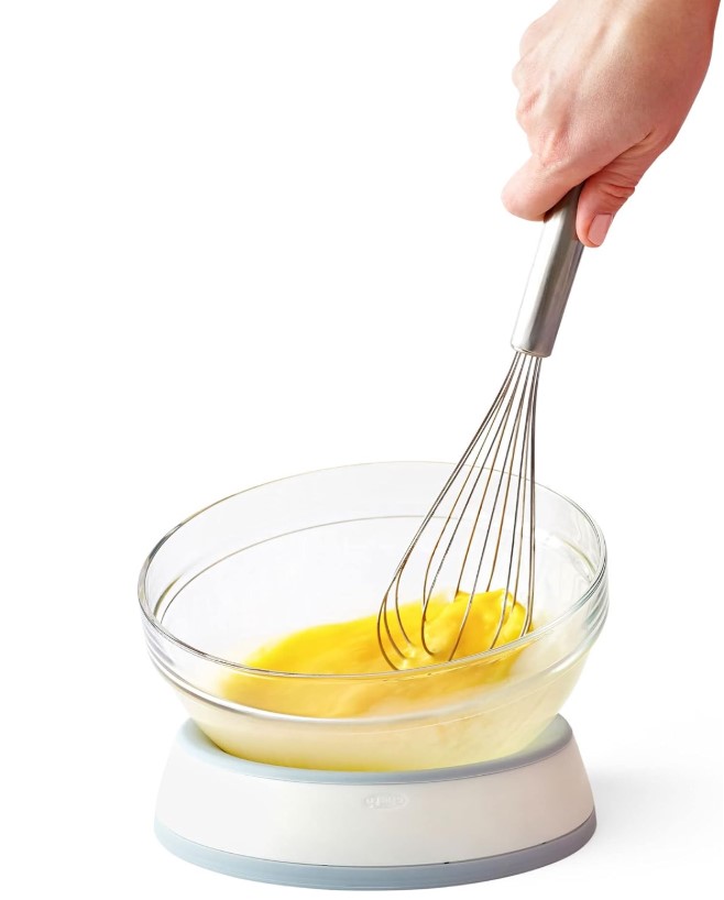 hand holding a whisk, stirring an egg in a glass bowl supported by a white and gray plastic circular cradle device
