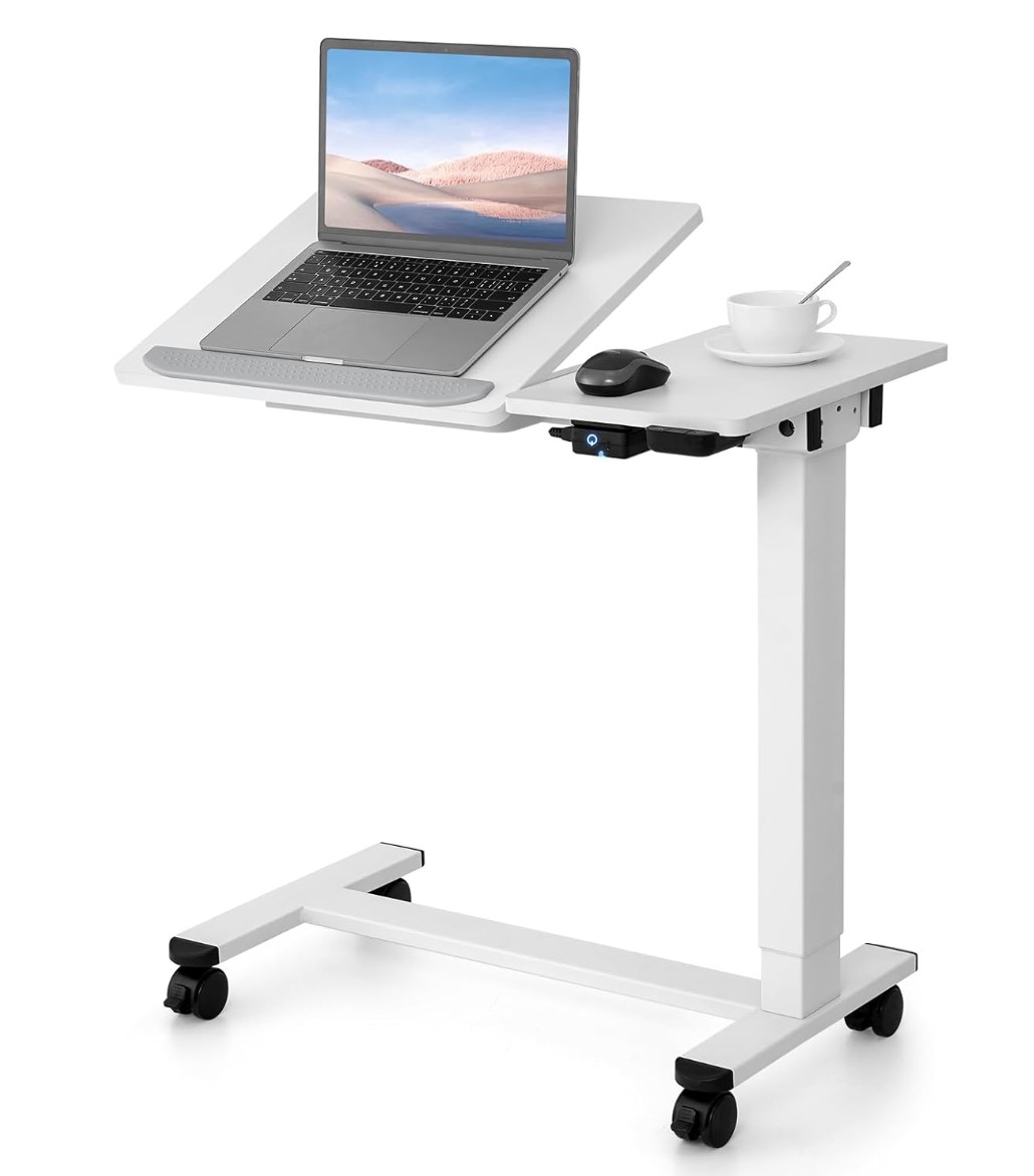 White height adjustable table on wheels with split top and left side of the table raised at an angle with a laptop sitting on it and right side of table is flat with a wireless mouse and cup on it