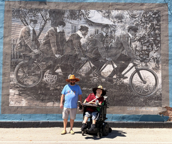 Photo of Michael in front of a mural of five men riding bikes. 